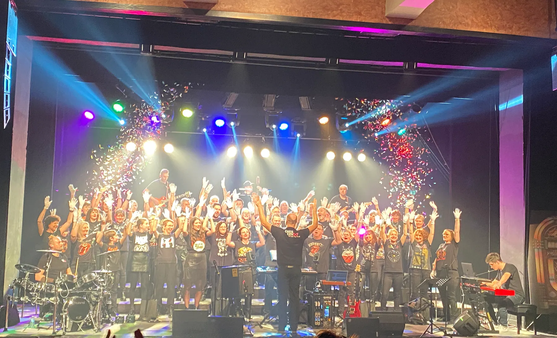 Le chœur au complet sur scène sous les projecteurs colorés, accompagné de ses musiciens